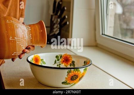 Weibliche Hand gießt Milch aus dem Keramikschüssel in eine Metallschüssel. In weißer Emaille lackiert, eine braune Kanne aus Steingut. Stockfoto