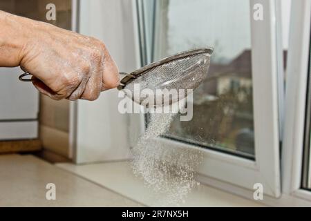 Weibliche Hand, die Mehl durch ein Handsieb für Mehl auf einem weißen Küchentisch gesiebt hat, Nahkopie. Ich koche Teig. Küchengerät aus Metall. Stockfoto