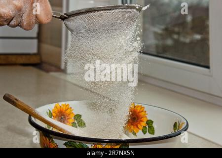 Weibliches Mehl, das von Hand durch ein manuelles Mehlsieb in einer Schüssel gesiebt wird, Nahkopie. Ich koche Teig. Küchengerät aus Metall. Stockfoto