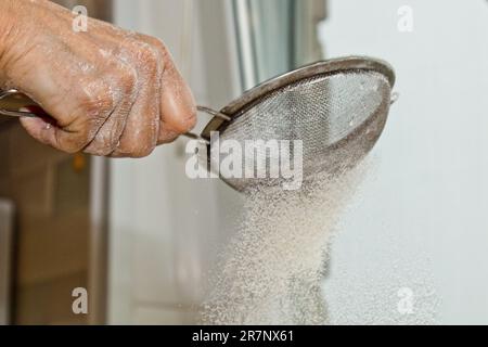 Weibliche Hand, die Mehl durch ein Handsieb für Mehl auf einem weißen Küchentisch gesiebt hat, Nahkopie. Ich koche Teig. Küchengerät aus Metall. Stockfoto