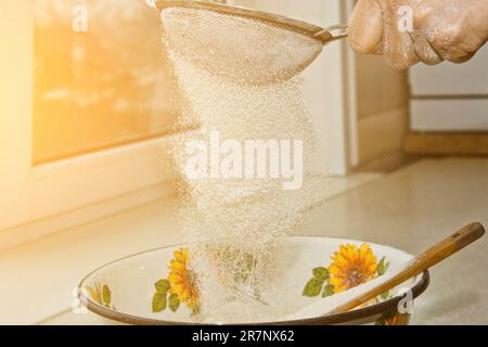 Weibliches Mehl, das von Hand durch ein manuelles Mehlsieb in einer Schüssel gesiebt wird, Nahkopie. Ich koche Teig. Küchengerät aus Metall. Stockfoto
