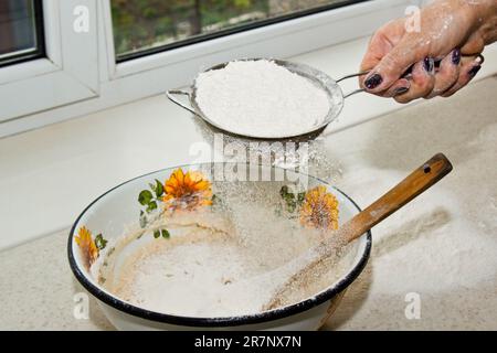 Weibliches Mehl, das von Hand durch ein manuelles Mehlsieb in einer Schüssel gesiebt wird, Nahkopie. Ich koche Teig. Küchengerät aus Metall. Stockfoto