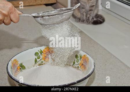 Weibliches Mehl, das von Hand durch ein manuelles Mehlsieb in einer Schüssel gesiebt wird, Nahkopie. Ich koche Teig. Küchengerät aus Metall. Stockfoto