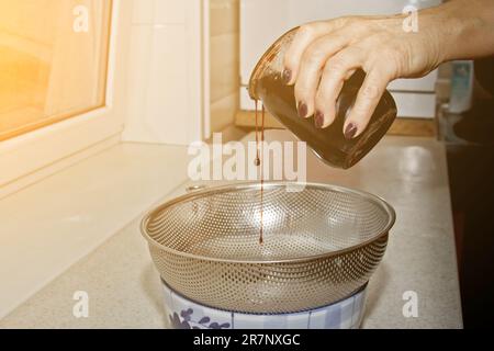 Weibliche Hände gossen Marmelade aus einem Glas durch ein Sieb vor dem Hintergrund des Kücheninnenraums in eine Schüssel. Backen, Kochen Stockfoto