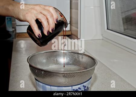 Weibliche Hände gossen Marmelade aus einem Glas durch ein Sieb vor dem Hintergrund des Kücheninnenraums in eine Schüssel. Backen, Kochen Stockfoto