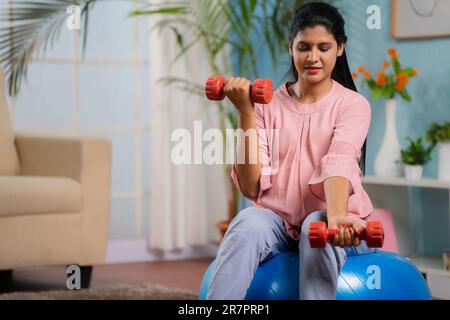 Indische schwangere Frau, die Sport mit den Kurzhanteln macht, während sie zu Hause auf dem Fitnessball sitzt - Konzept der aktiven Schwangerschaft, gesunden Lebensstil und Stockfoto