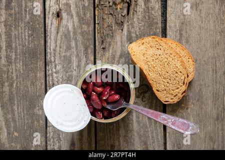 Rote Bohnen in einem eisernen Glas auf dem Tisch in der Küche zum Mittagessen neben einer Gabel und einer Scheibe Brot Stockfoto