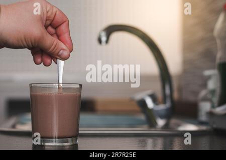 Nahaufnahme eines nicht wiedererkennbaren männlichen Weißen, der Milch und Kakao in einem Glas mit einem Löffel über einer Küchenbank umrührt. Stockfoto