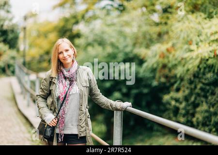 Außenporträt einer wunderschönen blonden Frau, die draußen posiert, einen khaki-Parka und einen rosa Schal trägt Stockfoto