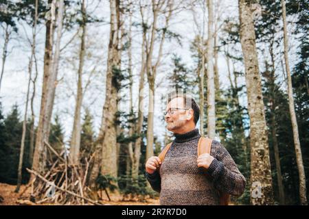 Ein hübscher Mann mittleren Alters, der im Wald wandert, einen Pullover, einen Rucksack und eine Brille trägt Stockfoto