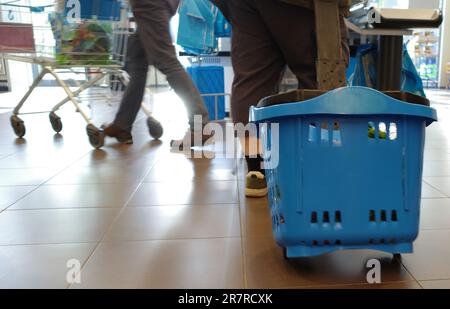 Die Beine eines Mannes, der einkauft. Er zieht einen blauen Plastikkorb auf Rollen hinter sich. Im Hintergrund sind die Beine einer Person mit einem SH Stockfoto