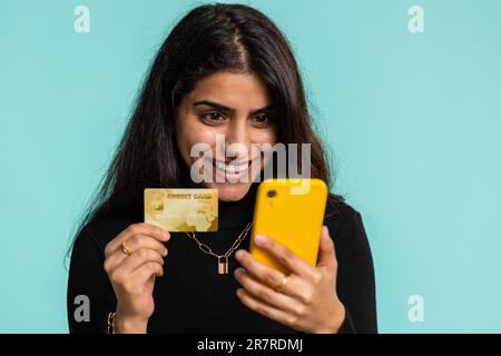 Ehrliche, indische Kundin, die bei Geldüberweisungen, Einkäufen im Internet und Bezahlung eine Kreditkarte und ein Smartphone verwendet. Finanzen und Internet. Hübsches Mädchen, isoliert auf blauem Hintergrund Stockfoto
