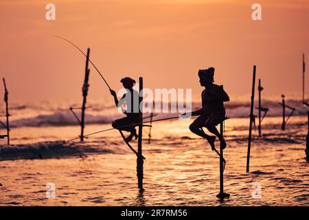 Silhouetten zweier traditioneller Fischer gegen das Meer in der Dämmerung. Traditionelles Pfahlfischen in der Nähe von Galle in Sri Lanka. Stockfoto