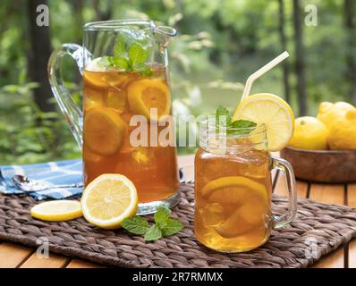 Eistee im Glasgefäß und Krug mit Zitrone und Minze im Freien auf einem Holztisch mit Naturhintergrund Stockfoto