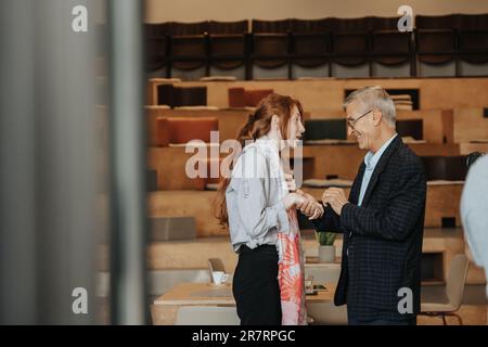 70s Firmenchef begrüßt seine junge, rothaarige Mitarbeiterin. Er schüttelt ihr die Hand Stockfoto