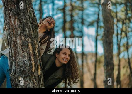 Zwei hübsche Mädchen, die lachten, sahen etwas Lustiges. Sie verstecken sich hinter einem Baum im Park Stockfoto