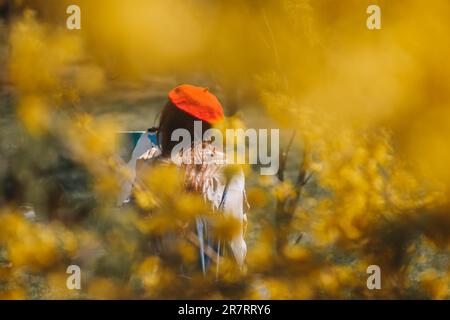 Eine Ingwer mit rotem Hut steht inmitten eines Feldes mit gelben Blumen und Gemälden, während sie im Park sitzt Stockfoto