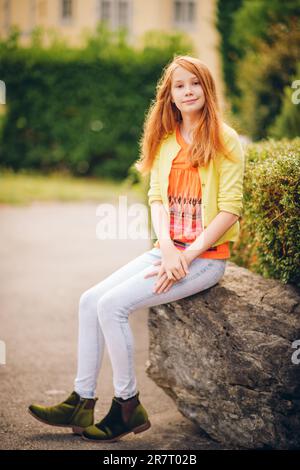 Außenporträt eines hübschen kleinen Mädchens mit rotem Haar, das ein orangefarbenes T-Shirt und eine gelbe Jacke trägt Stockfoto