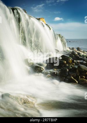 Ein majestätischer Wasserfall stürzt über einen steilen Felsvorsprung über den steinigen Strand Stockfoto