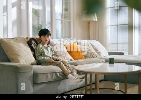 Ein kleiner asiatischer Junge, der zu Hause auf der Couch der Familie saß und traurig aussah Stockfoto