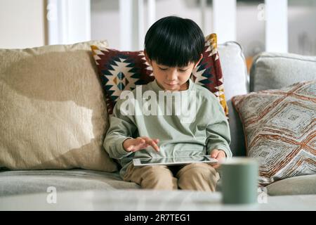 Fünf Jahre alter kleiner asiatischer Junge, der zu Hause auf dem Familientisch sitzt und einen digitalen Tablet-Computer benutzt Stockfoto