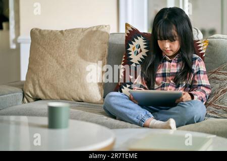 7-jähriges kleines asiatisches Mädchen, das zu Hause auf dem Familientisch sitzt und einen digitalen Tablet-Computer benutzt Stockfoto