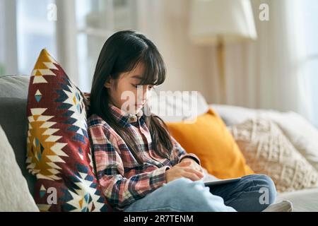 7-jähriges kleines asiatisches Mädchen, das zu Hause auf dem Familientisch sitzt und einen digitalen Tablet-Computer benutzt Stockfoto