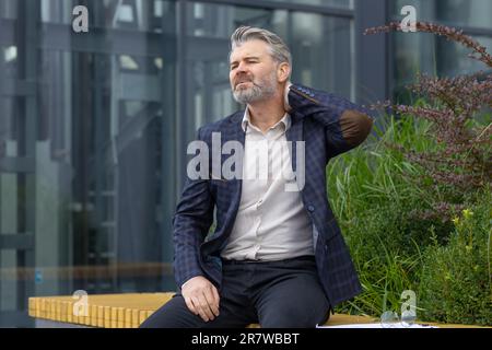 Übermüdeter, reifer Mann, der nach der Arbeit auf einer Bank vor dem Bürogebäude ruht, Geschäftsmann hat starke Nackenschmerzen, grauhaariger Boss im Anzug massiert Muskeln in der Mittagspause. Stockfoto