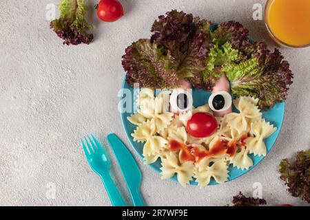 Fun Food für Kinder - lächelndes Gesicht von Pasta, Würstchen, Ei, Kirschtomaten und Salatblättern auf blauem Teller. Kreatives gesundes Frühstück für Kinder Stockfoto