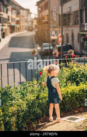 Außenporträt eines süßen, kleinen 3-4-jährigen Mädchens, das ein Denim-Schürzenkleid trägt und im öffentlichen Garten spielt Stockfoto