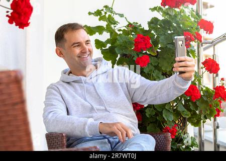 Ein lächelnder weißer Mann sitzt auf dem Stuhl auf der Terrasse, auf dem Balkon des neuen Campus, hält ein Smartphone, macht Selfie, spricht bei Videogesprächen. Glückliche weiße Männchen tragen Stockfoto