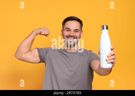 Glücklicher Mann, der die Thermoflasche hält und den Arm auf orangefarbenem Hintergrund zeigt Stockfoto