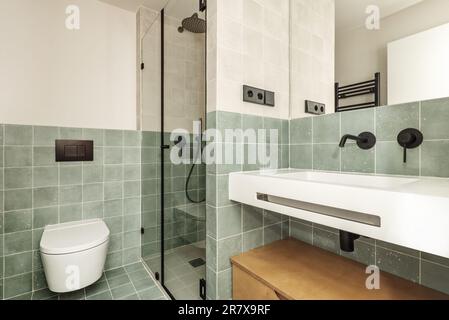 Badezimmer eines Hauses mit moderner Einrichtung aus kleinen grünen Fliesen, schwarzen Details in Wasserhähnen und Accessoires, rahmenlosem Spiegel und weißem Waschbecken aus Harz Stockfoto