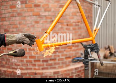 Eine nicht wiedererkennbare Person, die mit einem Lötbrenner die Farbe eines orangefarbenen Fahrradrahmens entfernt, der gerade in seiner Werkstatt renoviert wird. Stockfoto