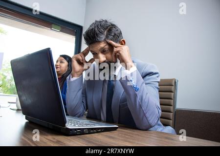 Gestresster und erschöpfter junger indischer Geschäftsmann sitzt im Büro und arbeitet an Laptop-Massage-Schläfen leiden an Kopfschmerzen.müde ungesunde männliche Angestellte str Stockfoto