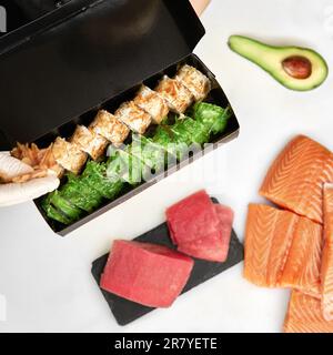 Sushi zum Mitnehmen. Draufsicht der Take-Away-Box mit Sushi-Rollen und Essstäbchen in schwarzer Papiertüte. Maki. Sashimi. Lachs. Thunfisch. Wach Auf. Asiatisch. Japanisch Stockfoto