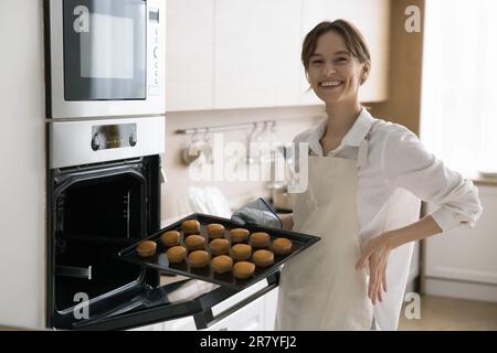 Junge Hausfrau in der Schürze, die ein Backblech mit Cupcakes hält Stockfoto