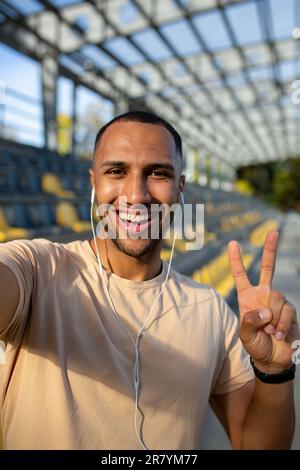 Nahaufnahme. Vertikales Foto. Junger afroamerikanischer Sportler steht im Stadion mit Kopfhörern, macht ein Selfie am Telefon, spricht bei einem Videoanruf. Zeigt der Kamera eine Supergeste an. Stockfoto