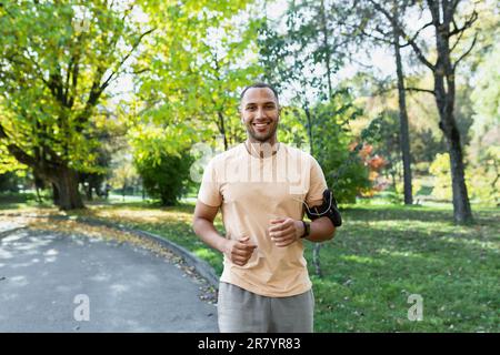 Lateinamerikanisch fröhlich und lächelnd im Park laufender Mann hört Musik und Audio-Podcasts, Läufer lächelt und schaut in die Kamera, fröhlicher Sportler draußen am Tag. Stockfoto