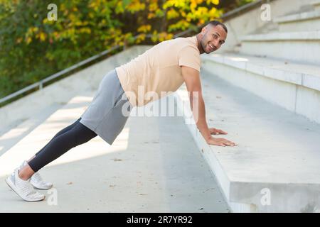 Erfolgreicher hispanischer Sportler, der verschiedene körperliche Übungen macht, morgens im Stadion trainiert, motiviert lächelt und in die Kamera schaut, Sportler in der Luft Fitness. Stockfoto