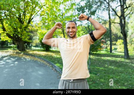 Porträt erfolgreicher Sportler zufrieden mit Trainingsergebnissen und Leistungen, schaute auf die Kamera und hielt die Hände in Triumph-Geste hoch, hispanischer Mann lief im Park mit Kopfhörern und Telefon. Stockfoto