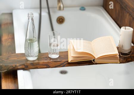 Ein Buch, eine Flasche Wasser, ein Glas und eine Kerze auf einem Holztablett über dem Badezimmer vor dem Hintergrund eines antiken Wasserhahns Stockfoto
