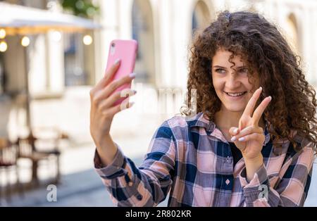 Hübsche junge Frau Bloggerin macht Selfie auf Smartphone, kommuniziert Videoanrufe online mit Abonnenten, nimmt Geschichten für soziale Medien Vlog im Freien auf. Ein Mädchen, das auf der sonnigen Stadtstraße spaziert Stockfoto