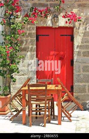 Wunderschöne Aussicht auf eine Gasse in Lefkada, mit Holztisch und 4 hölzernen Chaos umgeben, einer hölzernen roten Tür eines Steinhauses und Pflanzen. Stockfoto