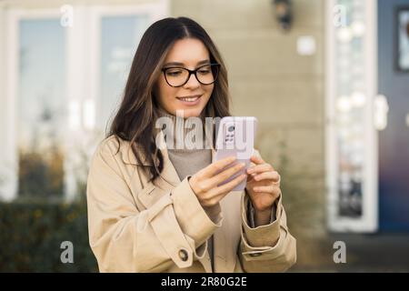 Lächelnde Frau mit Brille und beigefarbener Graben, die auf der Straße vor ihrer Veranda nach dem Telefon sucht. Stockfoto