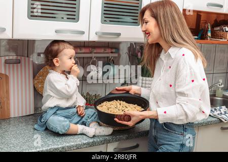 Mom und ihr einjähriges Mädchen in der Küche, mit Pasta in der Pfanne zum Abendessen Stockfoto