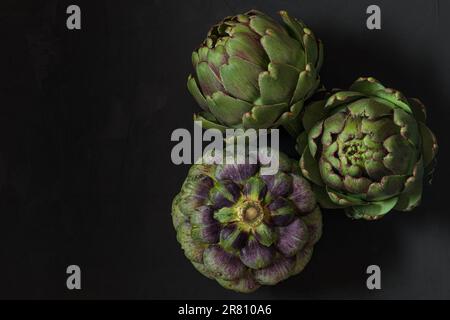 Frische reife Artischocken Blumen Gemüse auf schwarzem Hintergrund. Gesundes vegetarisches Essen. Draufsicht mit Kopierbereich Stockfoto