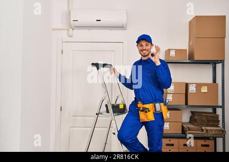 Junger spanischer Mann, der bei der Renovierung arbeitet und das Schild mit den Fingern gut macht, lächelnd freundlich und mit ausgezeichnetem Symbol Stockfoto