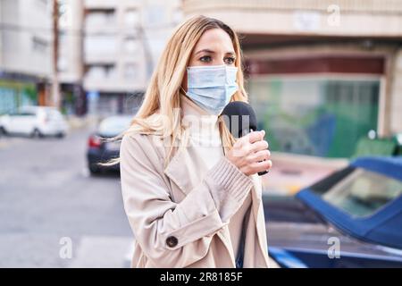 Junge blonde Reporterin trägt eine medizinische Maske und arbeitet auf der Straße Stockfoto