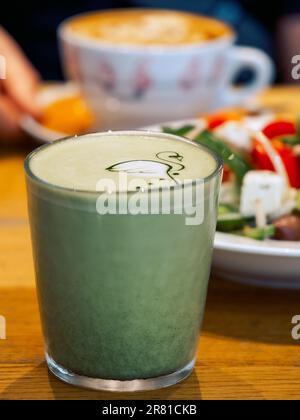 Heißer grüner Matcha-Tee in einem transparenten Glas mit Kunst, ein Teller mit griechischem Salat und eine Tasse Cappuccino-Kaffee in einem Restaurant. Stockfoto